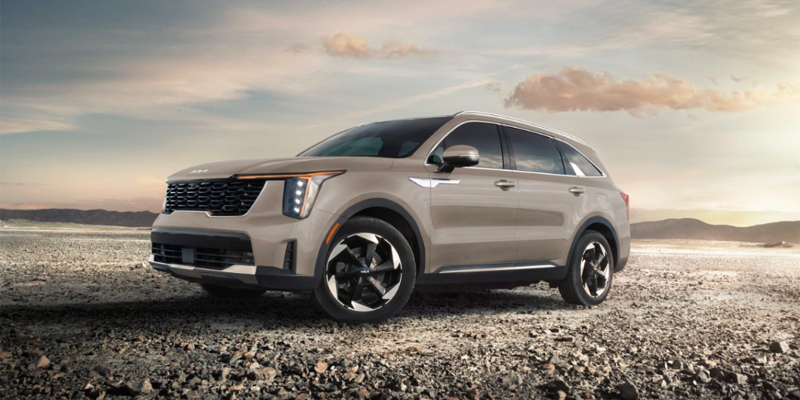 A stone colored 2025 Kia Sorento Plug-In Hybrid SUV parked on rocky terrain with the sun setting in the background and a partly cloudy sky overhead