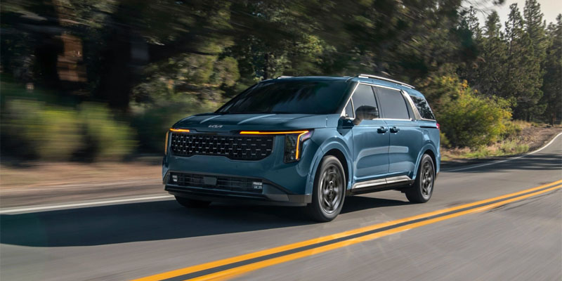 front driver's side view of a light blue 2025 Kia Carnival driving down a road surrounded by woods